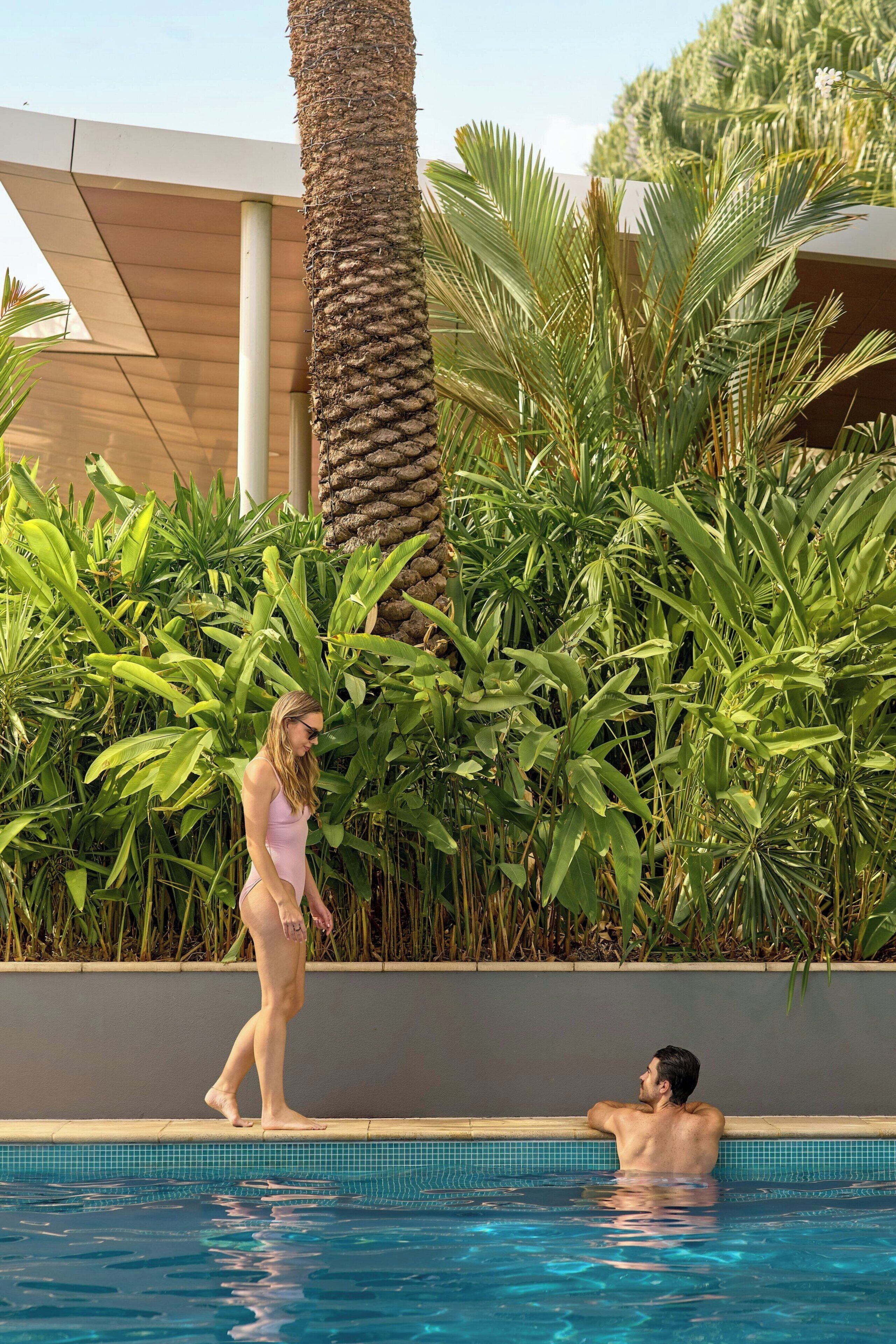 A swimming pool with clear blue water, surrounded by lush green tropical plants and a palm tree.