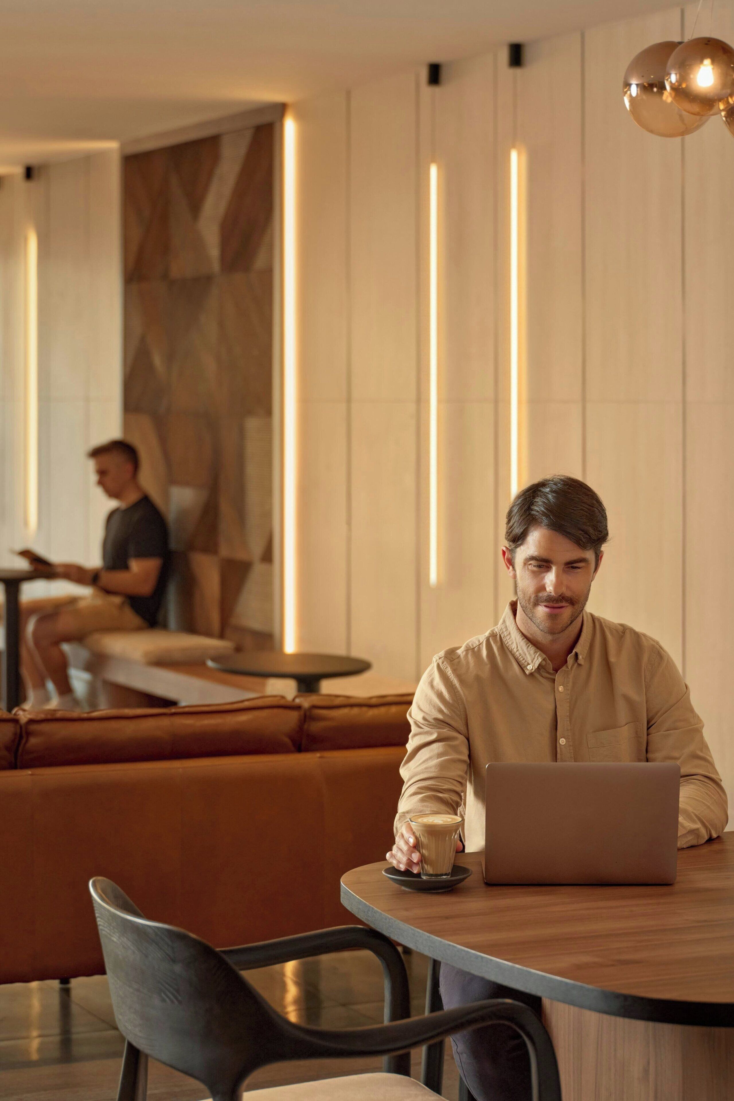Um homem sentado à mesa com um laptop e uma chávena de café num café moderno, com assentos de couro e iluminação pendente.