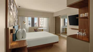 A hotel room with a large bed, a desk with a chair, a round table, a TV on a wooden shelf, and a view of the city through the windows.