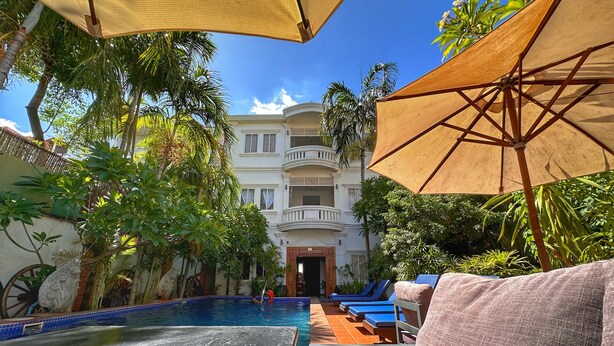 Un espace piscine agrémenté de chaises longues et de parasols, entouré d’une végétation luxuriante et bordé d’un bâtiment doté de balcons.