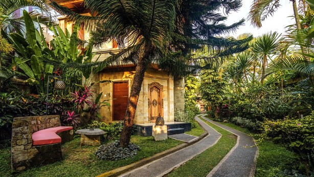A tropical garden with a stone pathway leading to a wooden door, surrounded by lush greenery and a red bench. - Rama Phala Resort & Spa (Ubud)