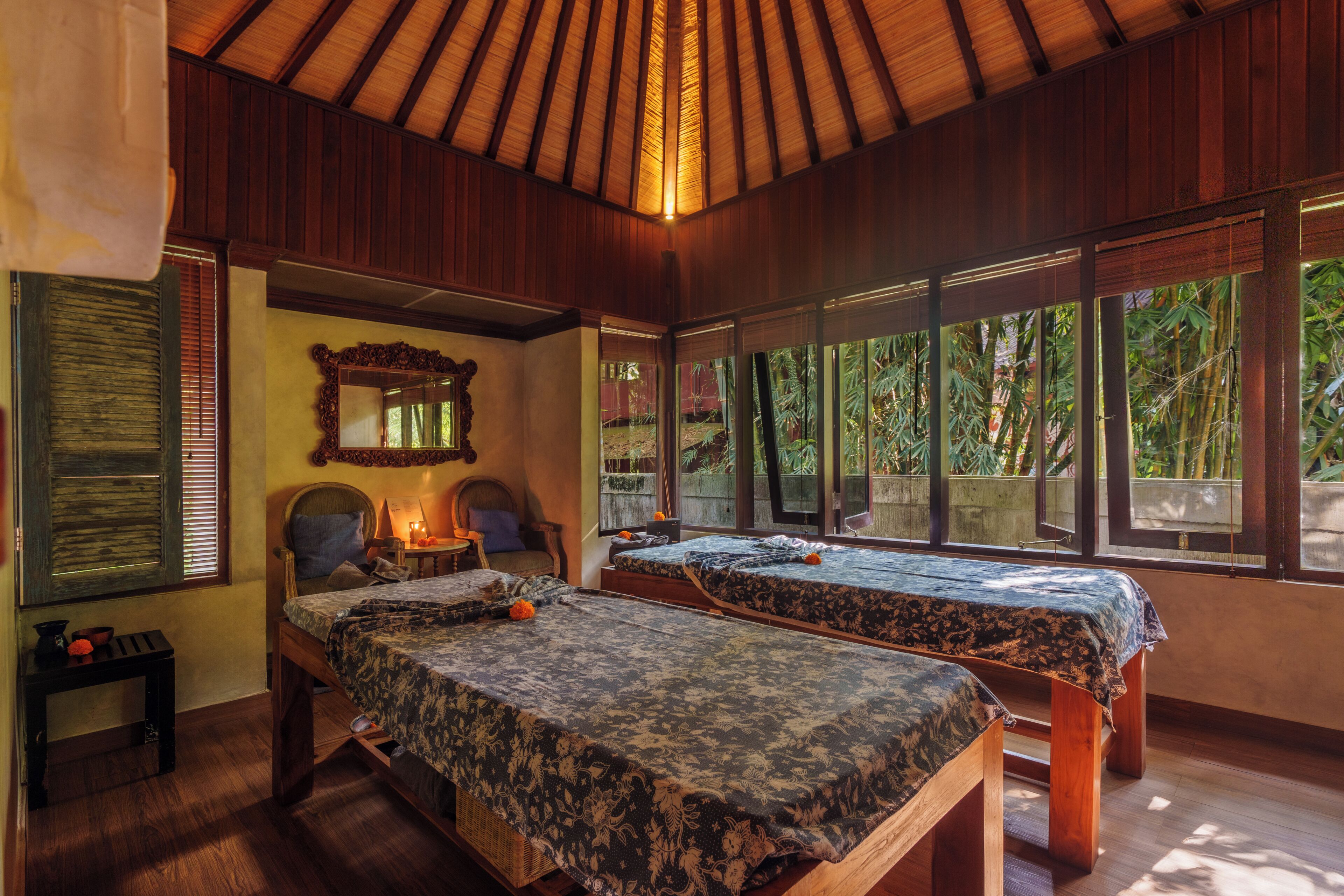 A room with two beds, a wooden ceiling, and large windows with a view of greenery.