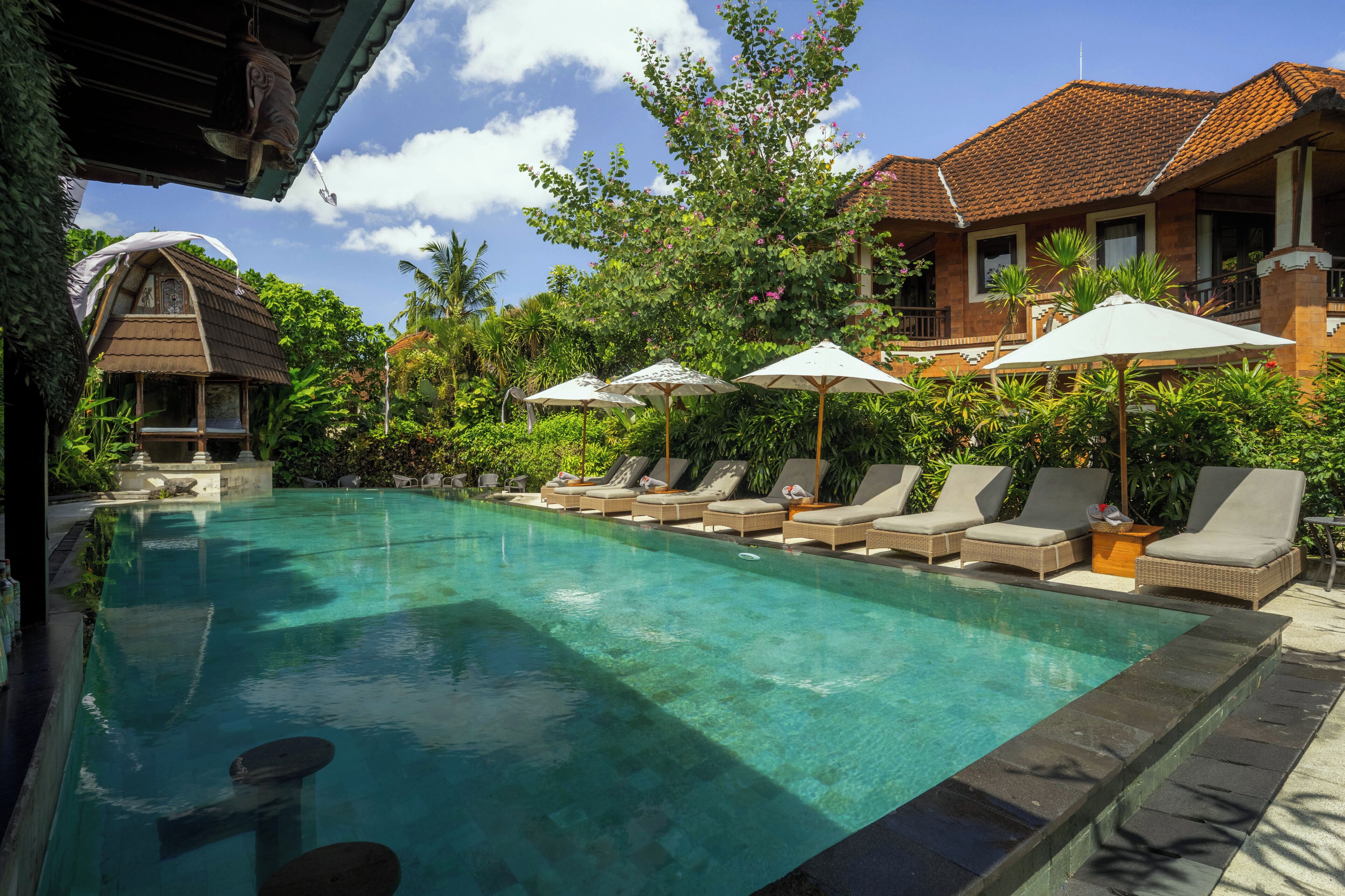 A clear swimming pool with lounge chairs and umbrellas, surrounded by lush greenery and traditional architecture.