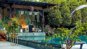 A covered outdoor seating area with a pool, surrounded by lush greenery and various plants.