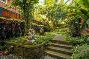 Una estatua de piedra de un mono con una flor roja en la cabeza, rodeada de exuberante vegetación y una escalera de piedra que conduce a una terraza de madera.