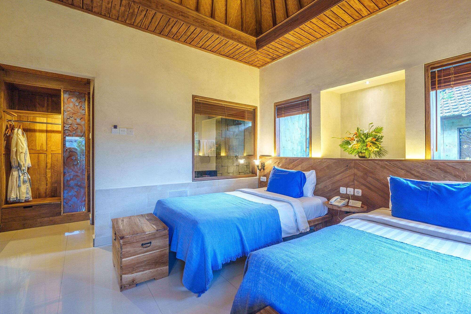 A hotel room with two beds, wooden headboards, a wooden wardrobe, a small wooden chest, and a mirror.