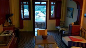 A compact room with a bed, a table, a chair, a small table with a bowl, and a view through a door to an outdoor area. - Bilios Apartments (Fournoi)