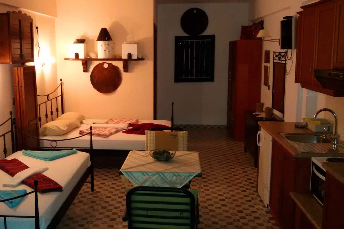 A small, compact room with a bed, a chair, a table with a bowl, a kitchen area with a sink, and a wall-mounted shelf with various items.