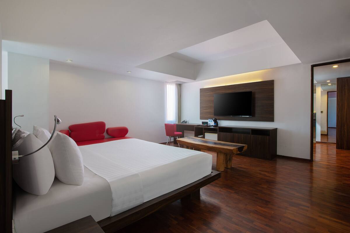 A modern hotel room with a large bed, a red chair, a wooden desk, and a flat-screen TV mounted on the wall.