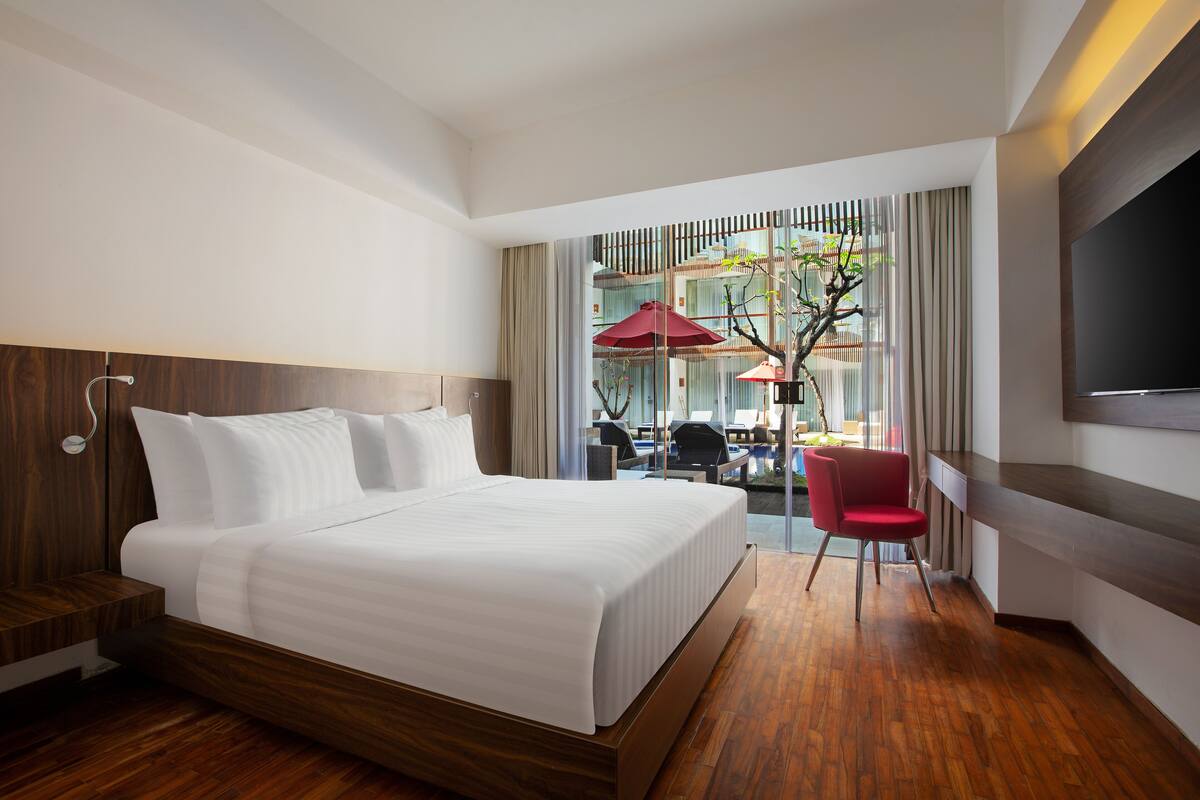 A modern hotel room with a large bed, wooden flooring, a TV, and a view of an outdoor seating area with umbrellas.