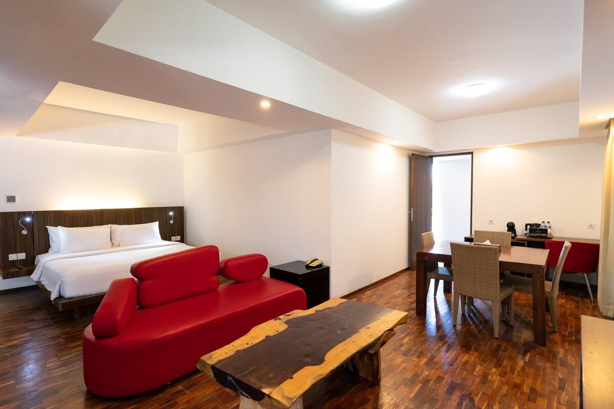 A modern hotel room with a bed, red sofa, wooden coffee table, dining table with chairs, and a small side table with a phone.