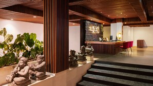 A reception area with a wooden reception desk, a stone statue of a seated figure, and a staircase leading to an upper level. - The Bene Hotel (Kuta)