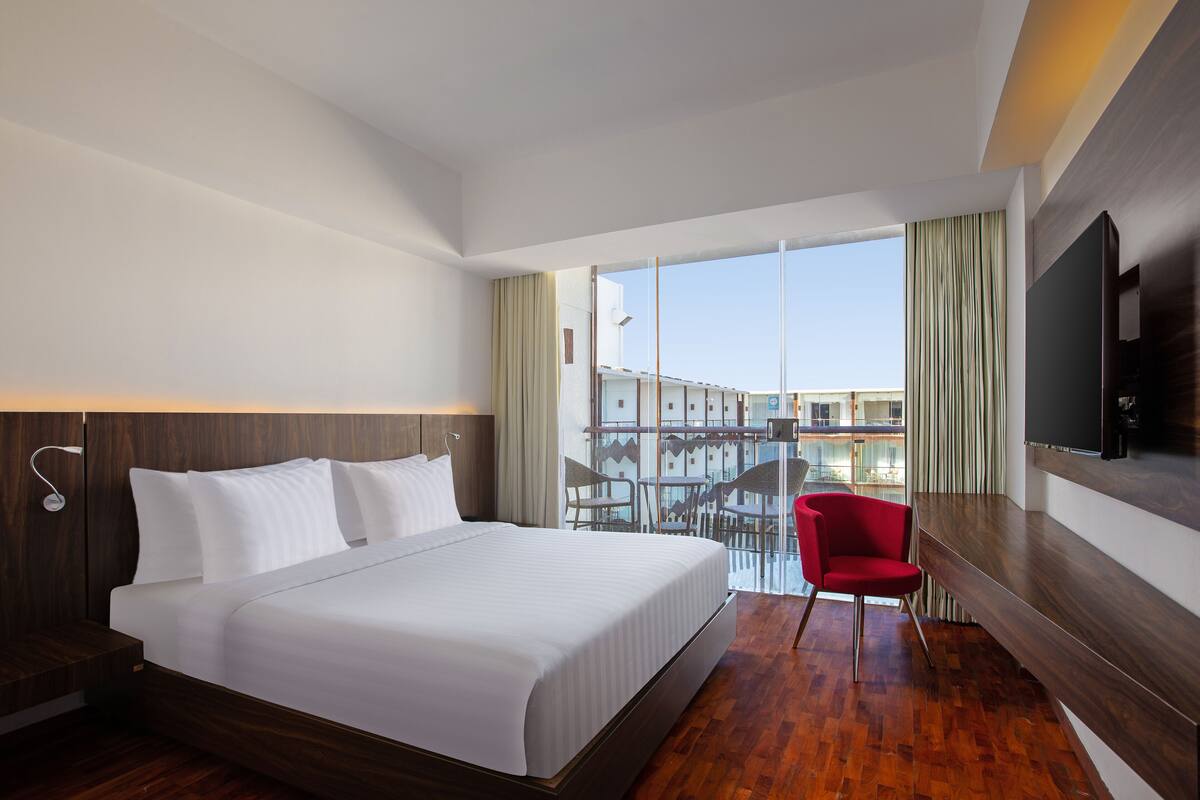 A hotel room with a large bed, a red chair, a TV, and a balcony with a view of buildings.
