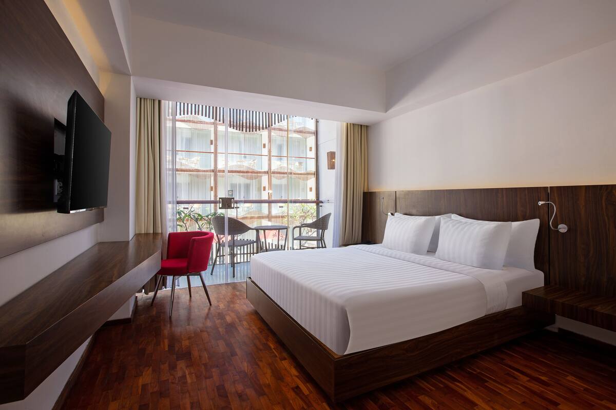 A modern hotel room with a large bed, a TV mounted on the wall, a small dining area with a table and chairs, and a view through large windows.