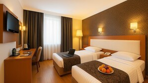 Quarto de hotel com duas camas, uma escrivaninha com um laptop, uma máquina de café e uma cesta de frutas na mesa de cabeceira.
