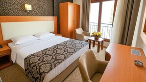 Quarto de hotel com cama grande, escrivaninha, cadeira, guarda-roupa e varanda com cortinas.
