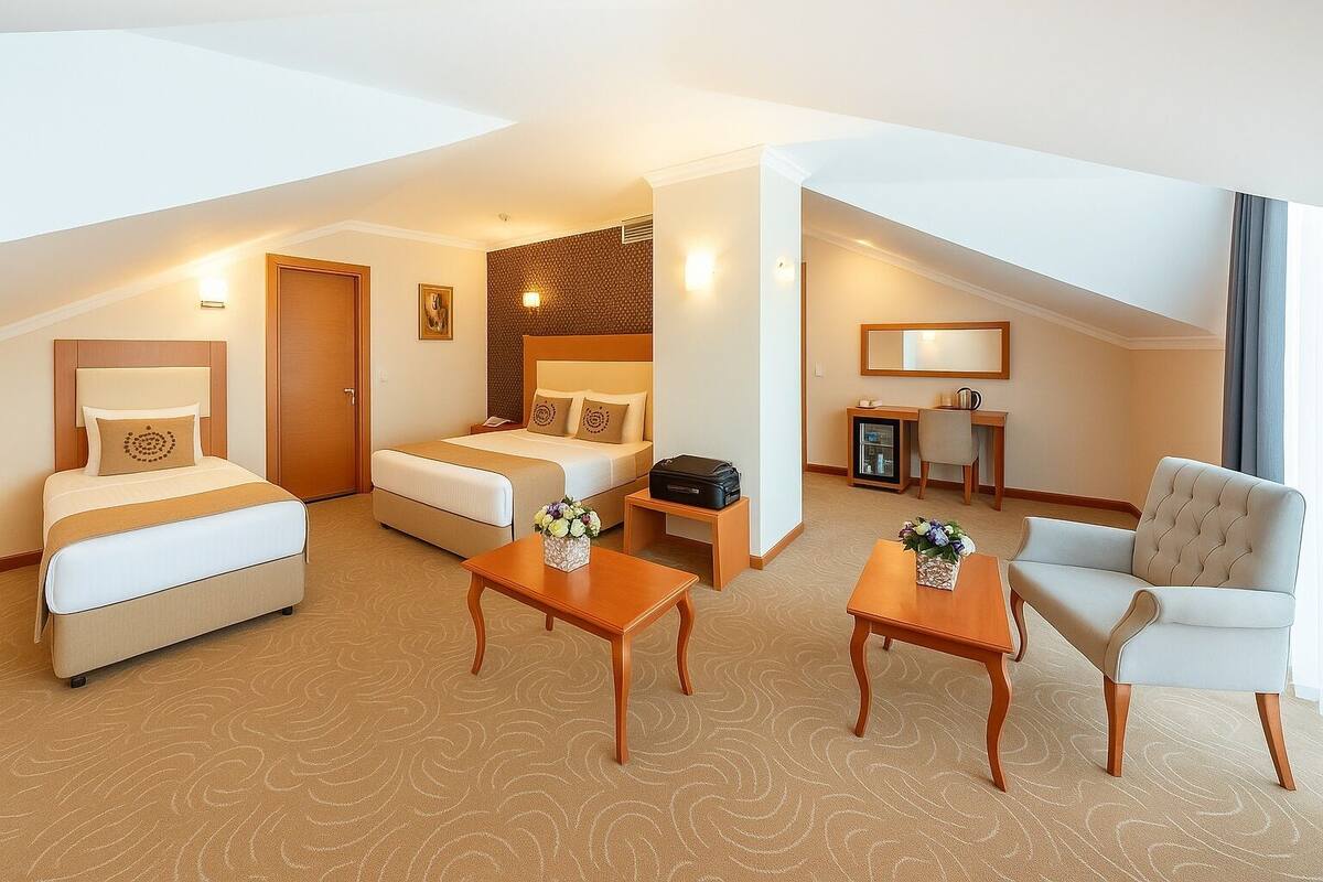 A hotel room with a bed, a sofa, a desk, and a chair. There is a small table with a vase of flowers. The room has a carpeted floor and a window with curtains.