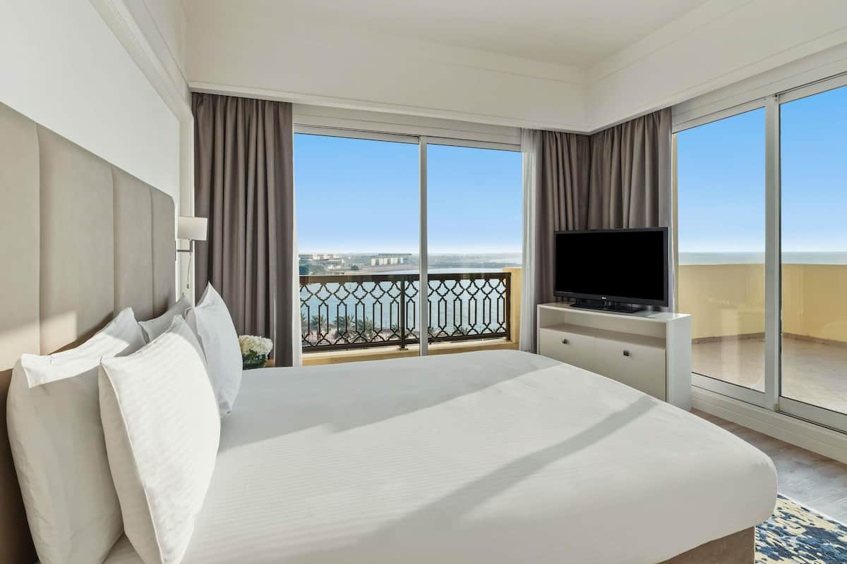 Habitación de hotel con una cama grande, televisor, balcón con vista al mar y ventana con cortinas.