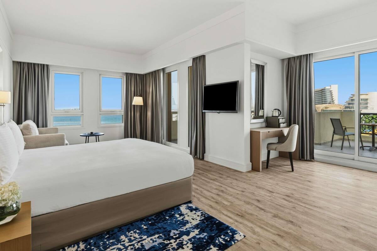 Habitación de hotel con una cama grande, un escritorio, un televisor y un balcón con vistas a la ciudad.