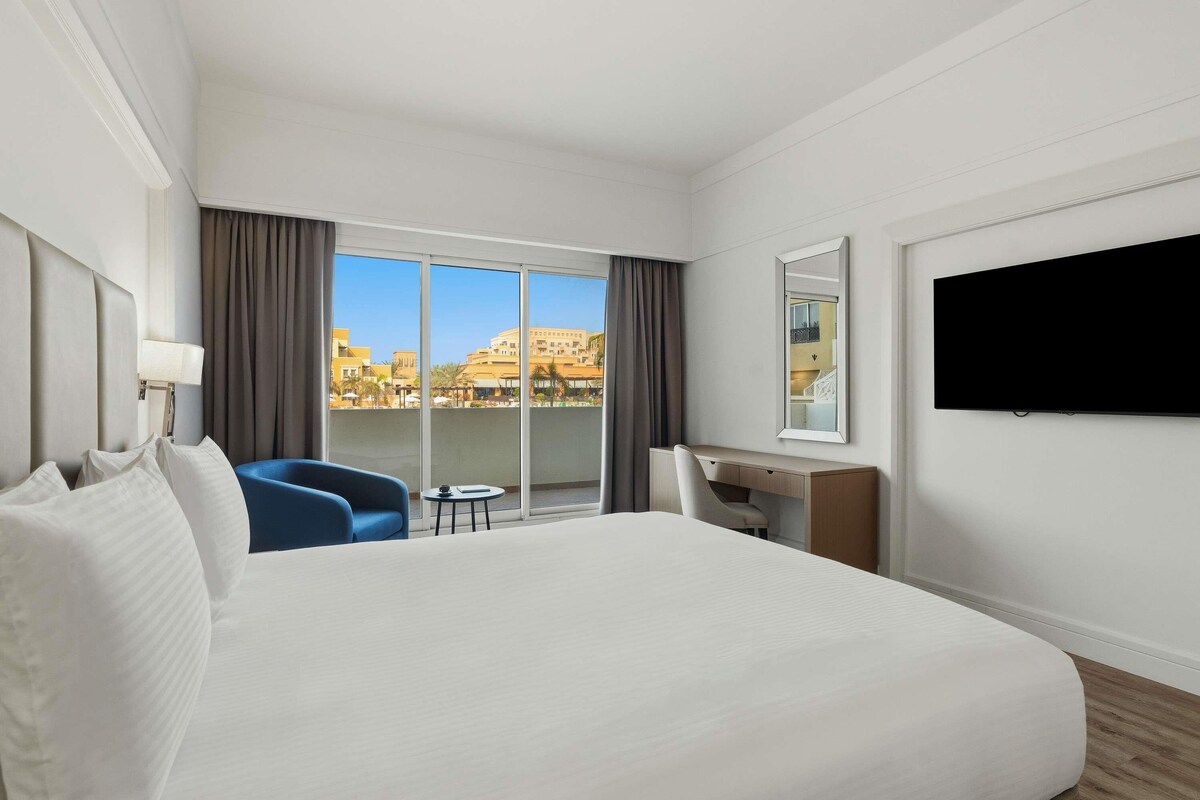 A hotel room with a large bed, a desk, a chair, a TV, and a view of buildings through a large window.