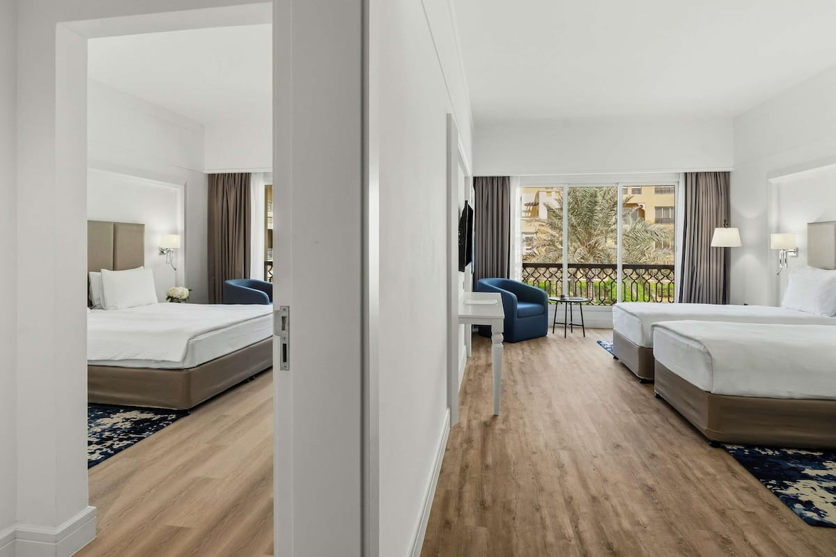 Habitación de hotel con dos camas, un escritorio, una silla y un balcón con vista a palmeras.