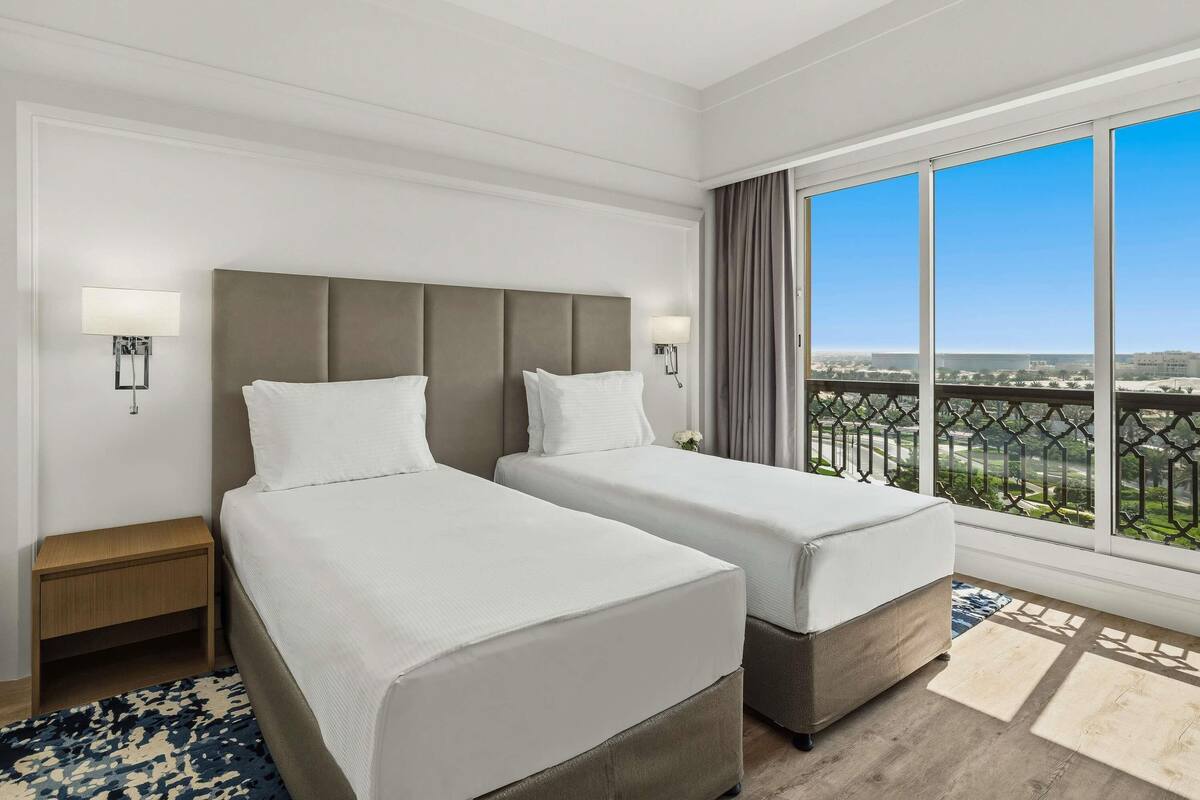 Habitación de hotel con dos camas, una mesita de noche, un ventanal con vistas y una alfombra estampada.