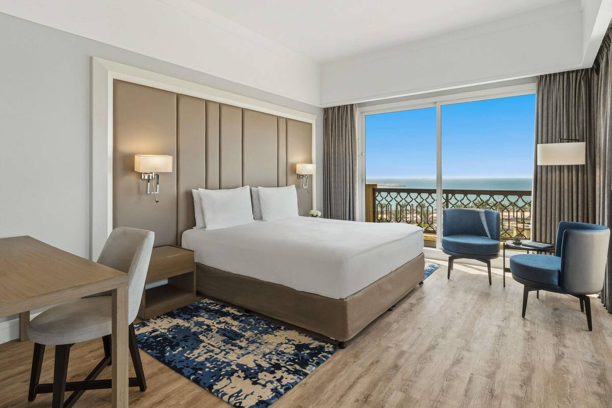 Habitación de hotel con una cama grande, un escritorio, dos sillas y vista al océano.