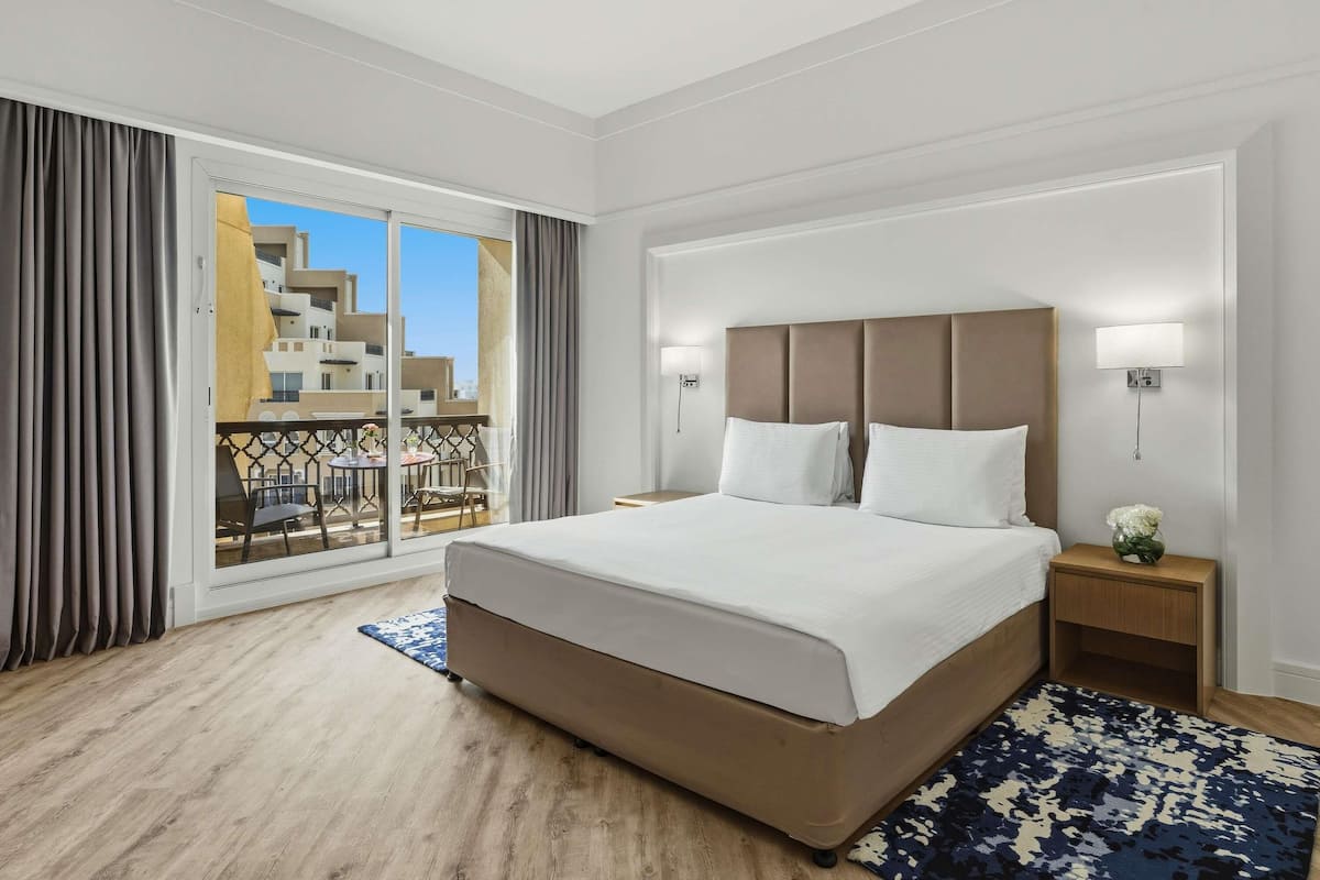Habitación de hotel con una cama grande, una mesita de noche, una lámpara de pared y vistas al balcón exterior.