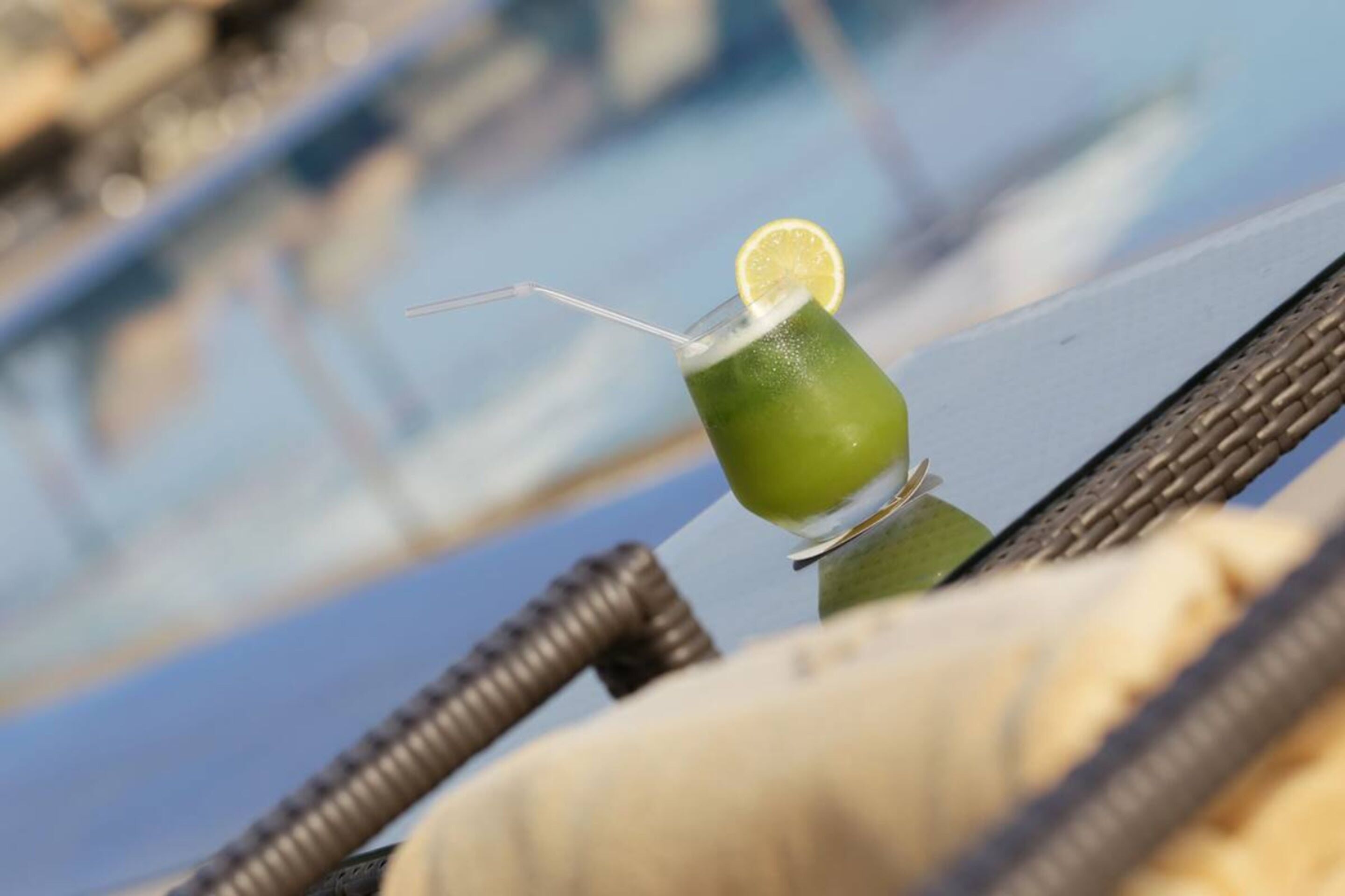 Un cocktail verde guarnito con una fetta di limone su un bicchiere, appoggiato su una sedia in vimini vicino a una piscina.