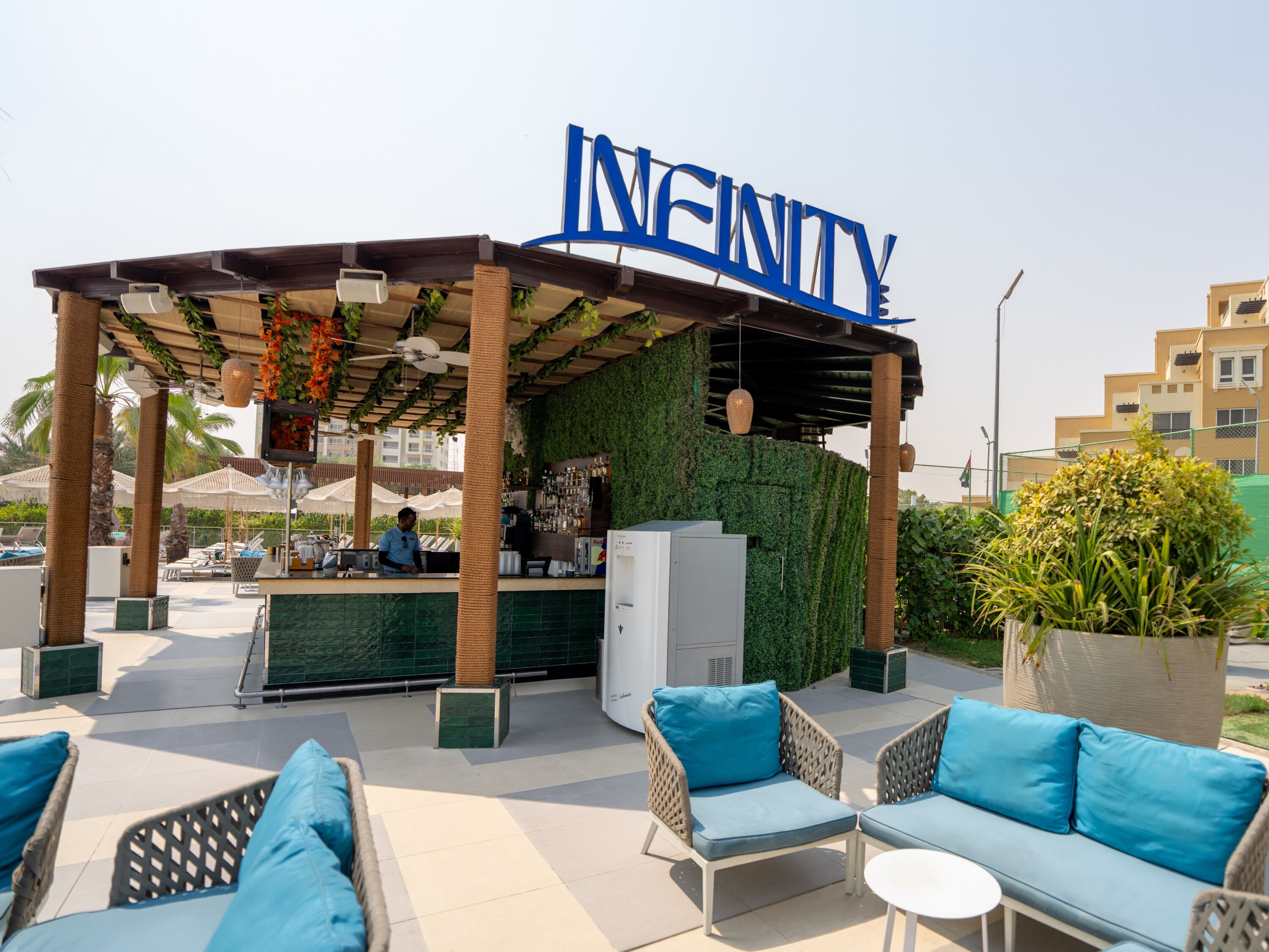 Un bar al terrat amb un cartell que diu "INFINITY", seients exteriors amb coixins blaus i una paret verda amb plantes.