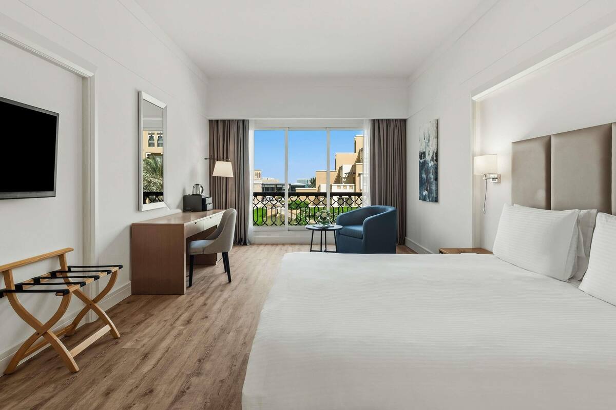 Una habitación de hotel con una cama grande, un escritorio, una silla, un televisor y un balcón con vistas a edificios.
