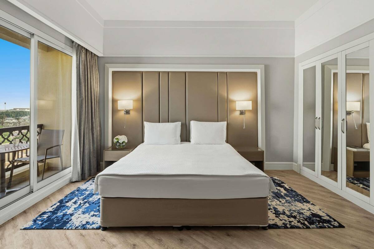 Habitación de hotel con una cama grande, dos lámparas de noche, vistas a través de un ventanal corredero y un balcón con mesa y sillas.