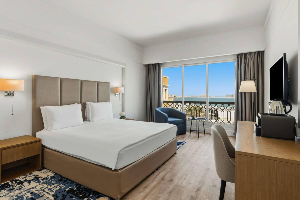 Habitación de hotel con una cama grande, un escritorio, una silla, un televisor y un balcón con vista al mar.