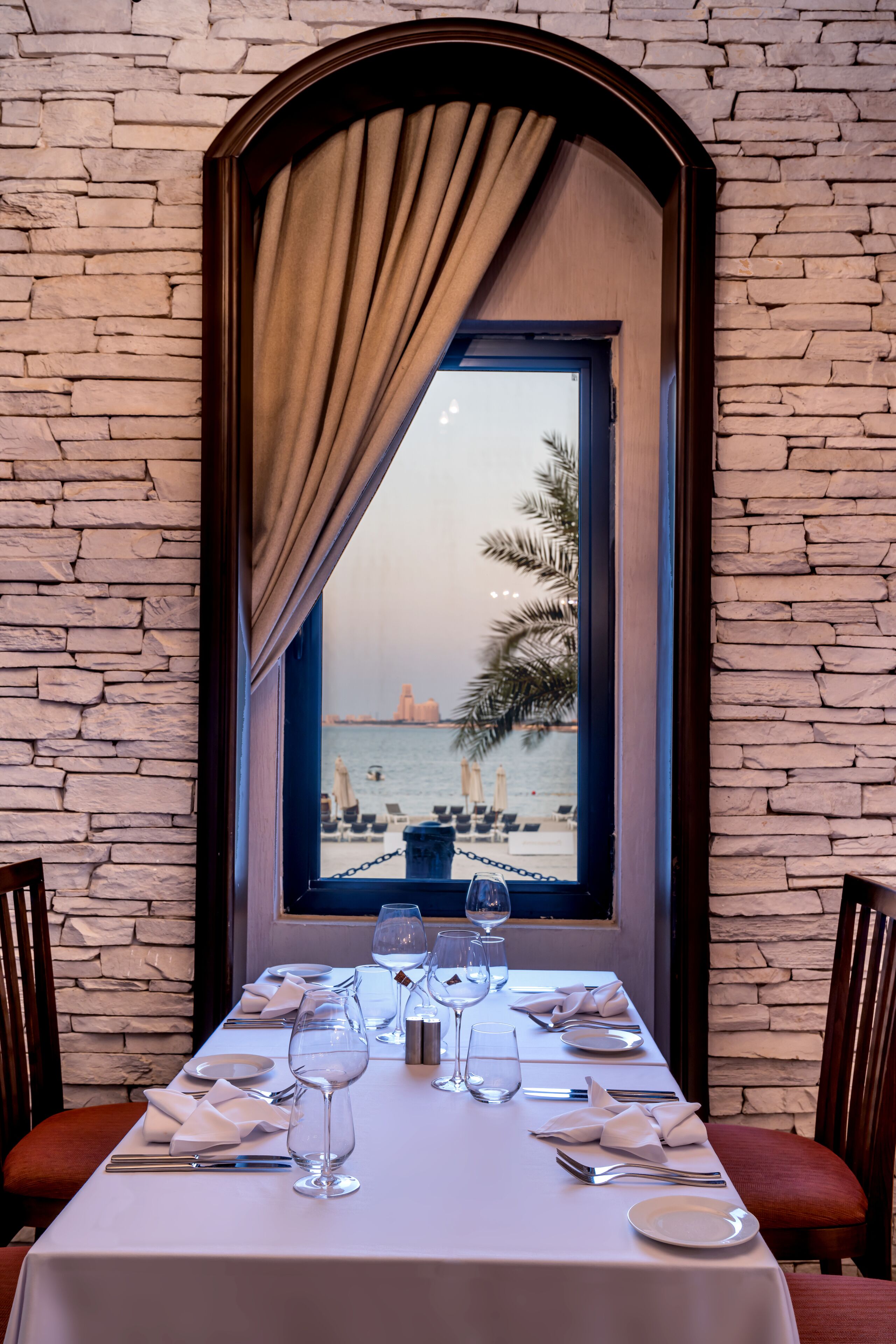 Une table à manger dressée avec de la vaisselle blanche et des verres à vin, donnant sur une plage bordée de palmiers et sur une vue lointaine de la ville.