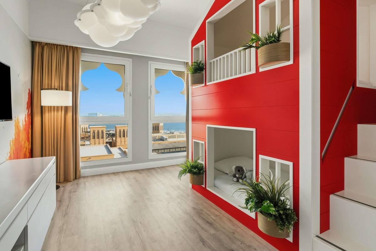 Una habitación de hotel moderna con una decoración en rojo y blanco, grandes ventanas con vistas a la ciudad, un candelabro en el techo y plantas en macetas.