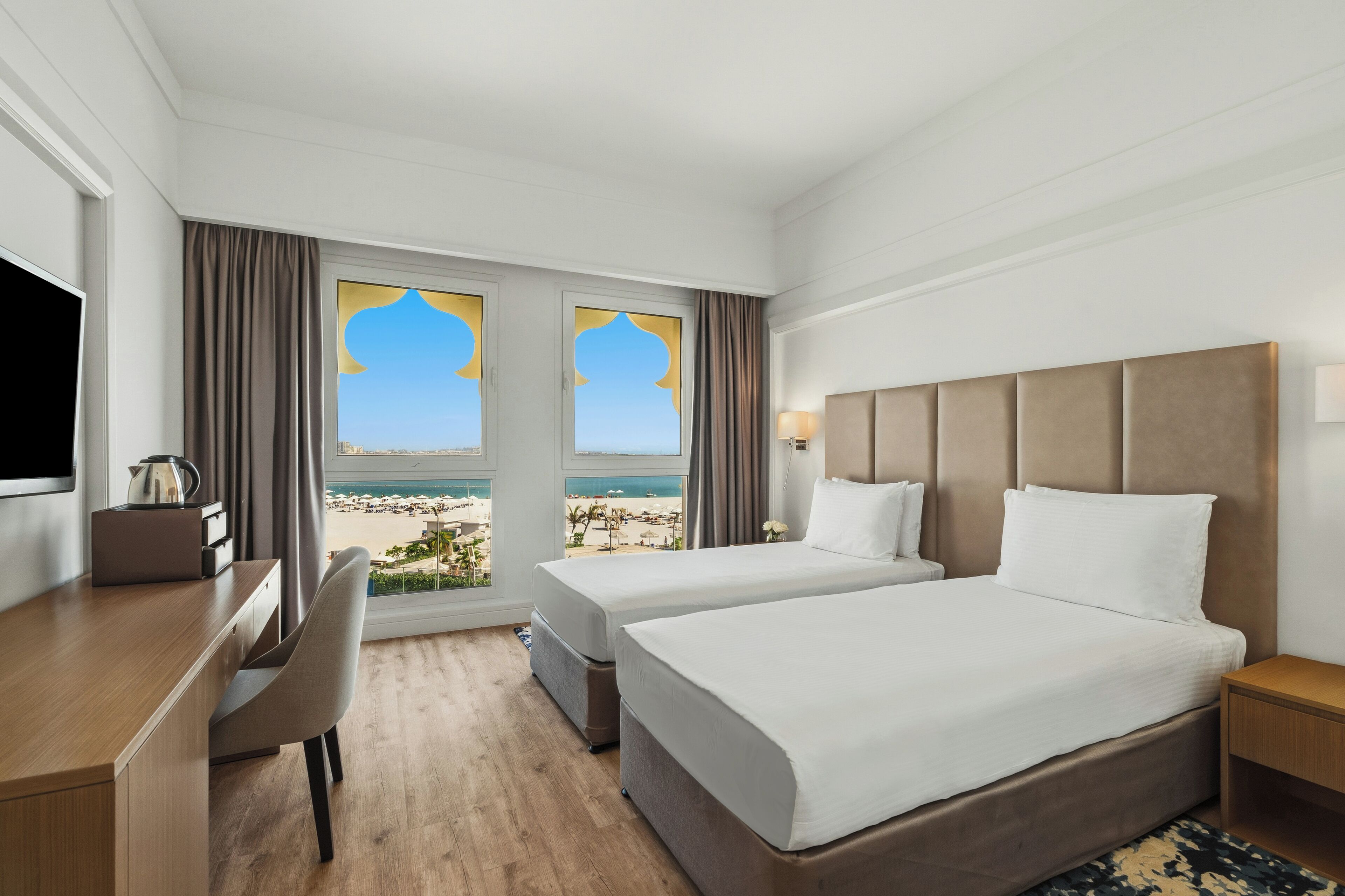 A hotel room with two beds, a desk, a chair, and a view of the beach.