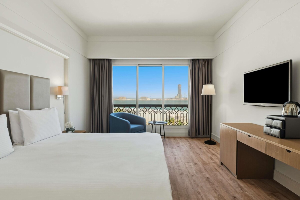 A hotel room with a bed, a desk, a TV, a chair, and a balcony with a view.