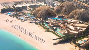 Vista aerea di un resort con diversi edifici, una grande area piscina e una spiaggia con lettini.