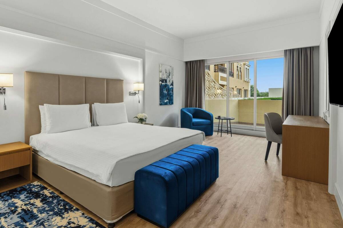 Habitación de hotel con una cama grande, un sillón azul, una mesita y vistas a edificios a través de la ventana.