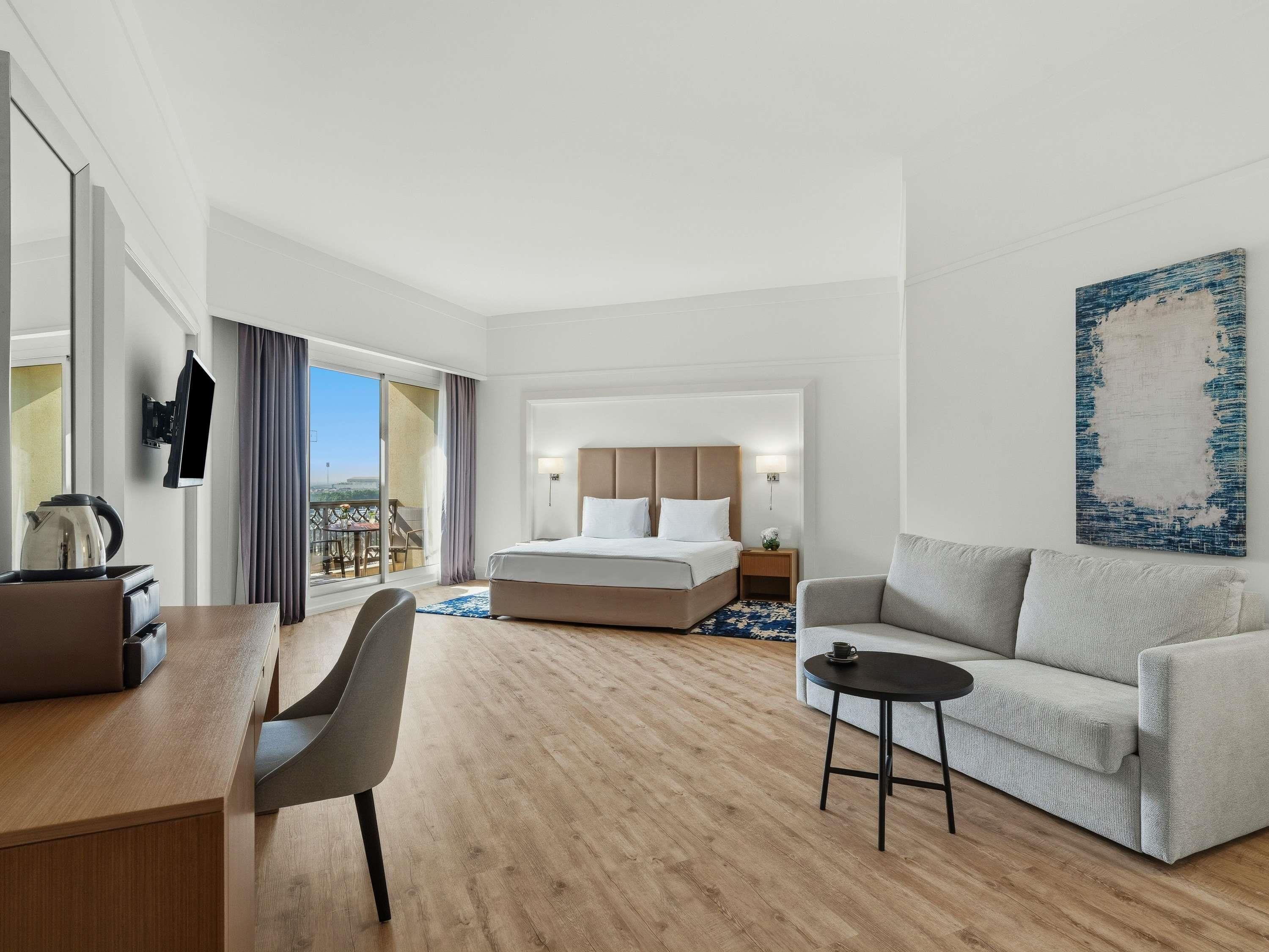 A hotel room with a bed, a desk, a chair, a sofa, a TV, and a painting.