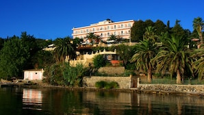A multi-story hotel with balconies, situated by a body of water, surrounded by greenery and palm trees. - Aegli Hotel (Corfu)