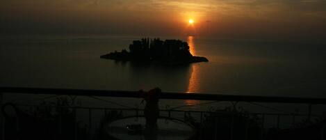 Vista al atardecer desde un balcón con mesa y sillas, con vista a un mar tranquilo y una pequeña isla.