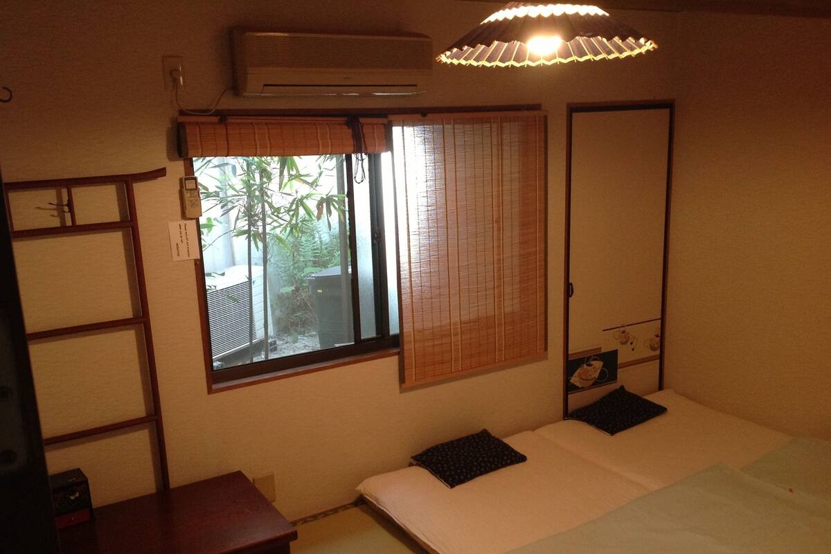 A bedroom with a bed, a bedside table, a wardrobe, a window with blinds, and an air conditioning unit.