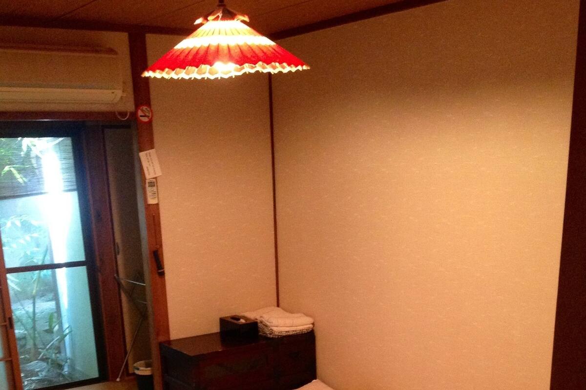 A small, neatly made bed with a red and white lamp above it, a wooden nightstand with a small item on it, and a door leading to a balcony.