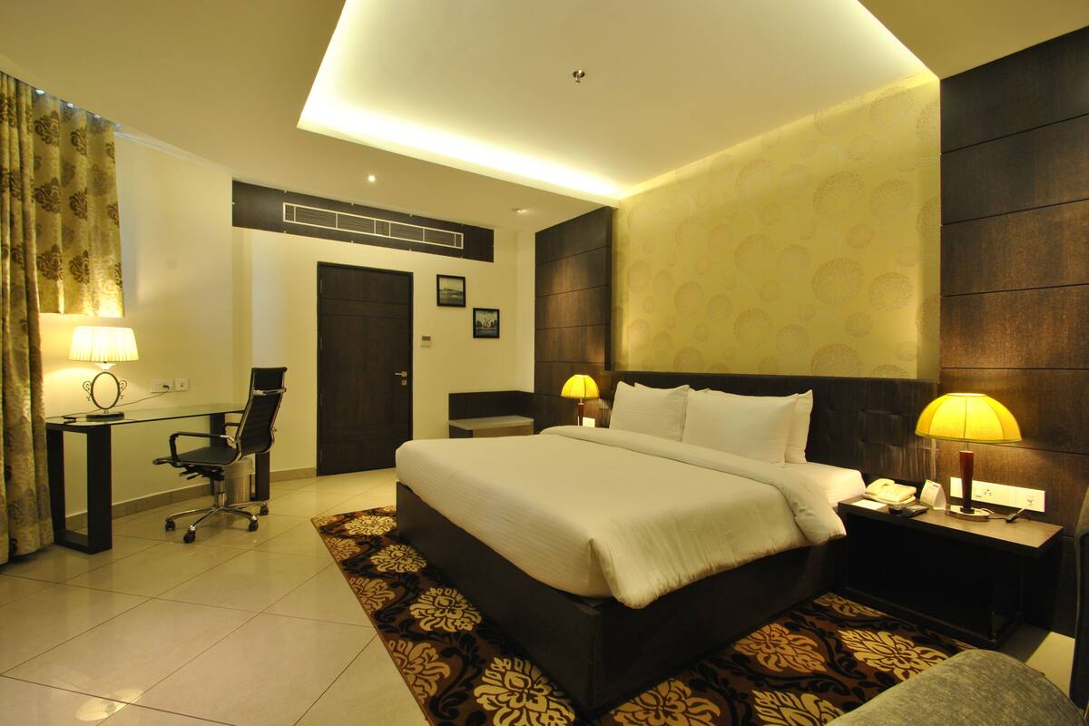 A hotel room with a large bed, two bedside lamps, a desk with a chair, a small table, and a patterned rug.