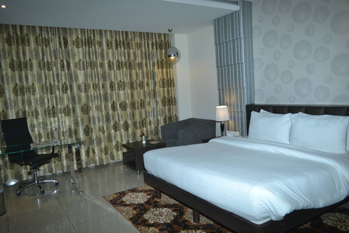 A hotel room with a large bed, a glass desk, a chair, a nightstand, and a lamp.