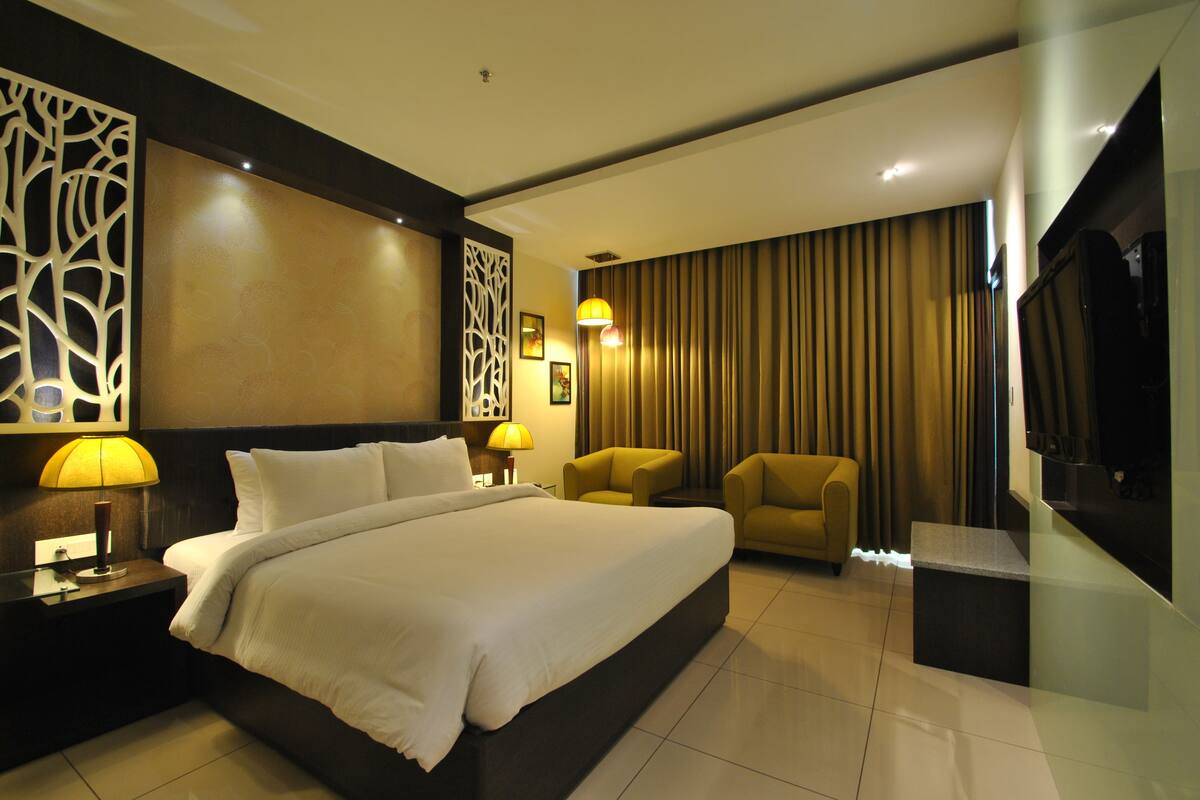 A modern hotel room with a large bed, two armchairs, a TV, and a decorative wall panel.