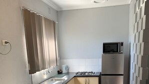 A compact kitchen with a sink, stove, microwave, and refrigerator. There is a dining table with two white chairs and a small potted plant. - Hello Adelaide Motel and Apartments (Frewville)