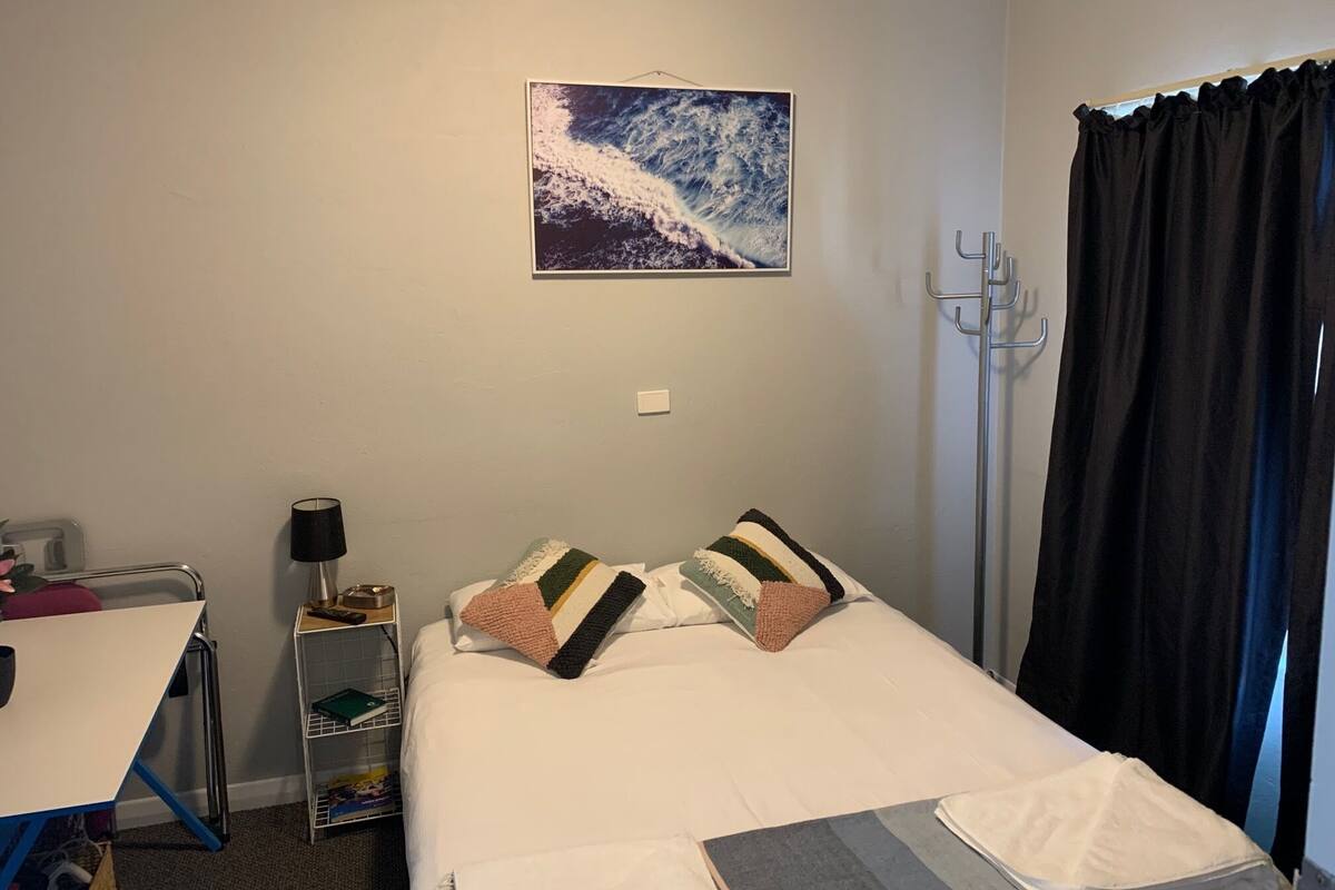 A hotel room with a bed, a nightstand, a lamp, a coat rack, and a window with curtains.