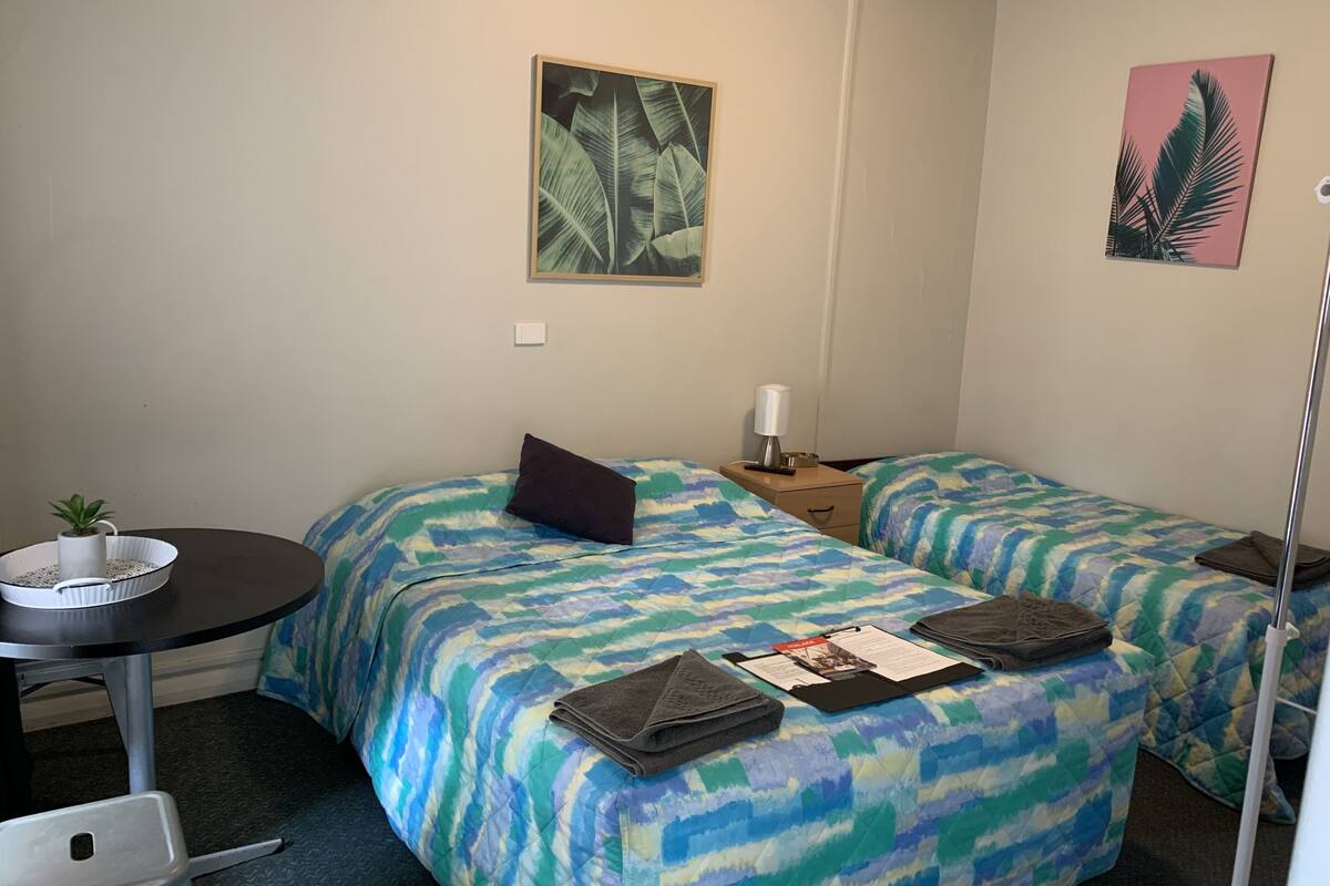 A hotel room with two beds, a small table with a plant, a nightstand with a lamp, and artwork on the walls.
